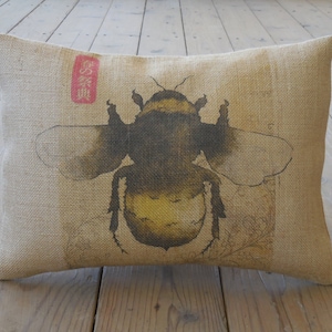 May include: A beige burlap pillow with a black and yellow bumblebee illustration. The bumblebee is surrounded by a brown and white floral design. The pillow has a red and white label with Japanese characters.