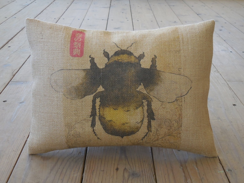 May include: A beige burlap pillow with a black and yellow bumblebee illustration. The bumblebee is surrounded by a brown and white floral design. The pillow has a red and white label with Japanese characters.