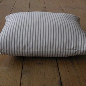 May include: A white pillow with thin grey stripes. The pillow is lying on a wooden floor.