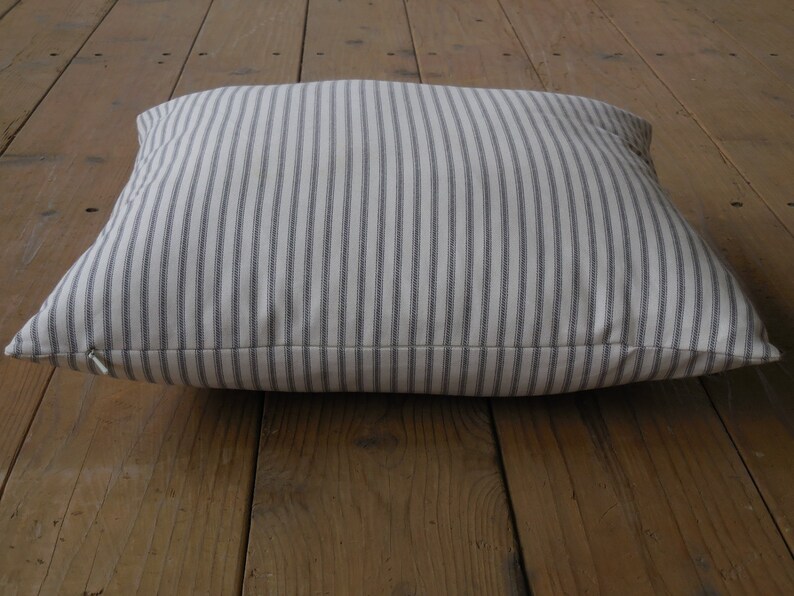 May include: A white pillow with thin grey stripes. The pillow is lying on a wooden floor.