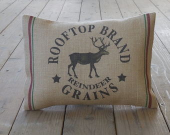 May include: A tan burlap pillow with red and green trim. The pillow has a black and white graphic of a reindeer with the text "Rooftop Brand Reindeer Grains" printed on it.