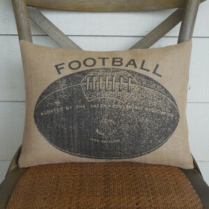 May include: A brown burlap pillow with the word "FOOTBALL" printed in black. A black and white illustration of a football is in the center of the pillow.