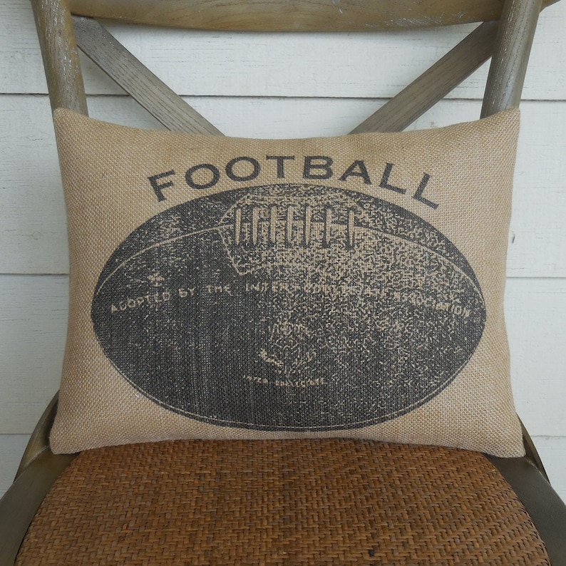 May include: A brown burlap pillow with the word "FOOTBALL" printed in black. A black and white illustration of a football is in the center of the pillow.