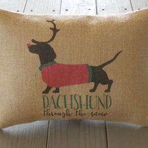 May include: A brown burlap pillow with a black and red dachshund wearing a red sweater and a red nose. The text "Dachshund through the snow" is printed on the pillow.