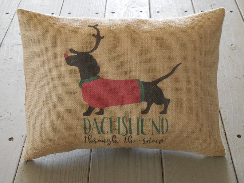 May include: A brown burlap pillow with a black and red dachshund wearing a red sweater and a red nose. The text "Dachshund through the snow" is printed on the pillow.