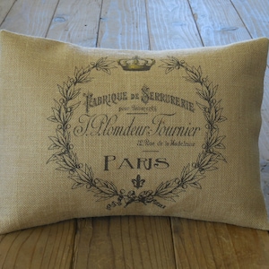 May include: A tan burlap pillow with a black and white design featuring a crown, a wreath of leaves, and the text "Fabrique de Serrurerie pour Industries J. Blondeau Fournier 12, Rue de la Madeleine PARIS".