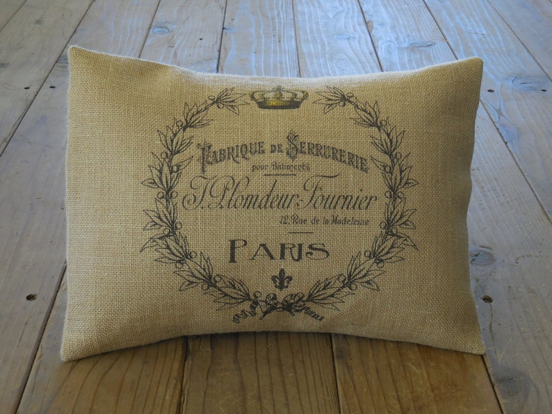 May include: A tan burlap pillow with a black and white design featuring a crown, a wreath of leaves, and the text "Fabrique de Serrurerie pour Industries J. Blondeau Fournier 12, Rue de la Madeleine PARIS".