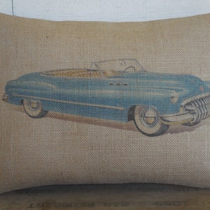 May include: A blue vintage convertible car printed on a burlap pillow. The pillow is a light brown color and has a rough texture.