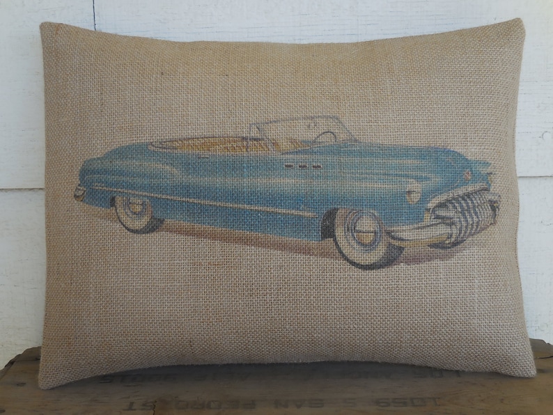 May include: A blue vintage convertible car printed on a burlap pillow. The pillow is a light brown color and has a rough texture.