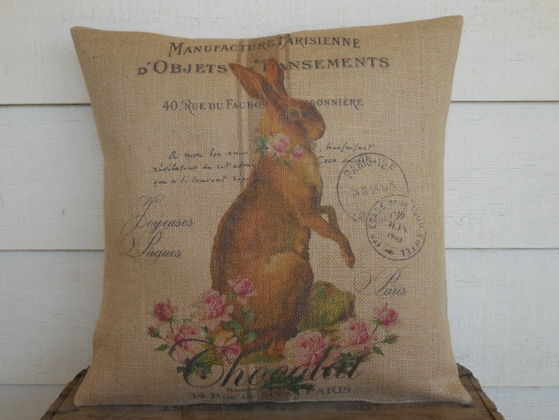 Może przedstawiać: Poduszka z juty z vintage'owym francuskim wzorem przedstawiającym brązowego kr&oacute;lika, r&oacute;żowe r&oacute;że i tekst "Manufacture Parisienne d'Objets d'Amusements 40, Rue du Faubourg Sonniere".