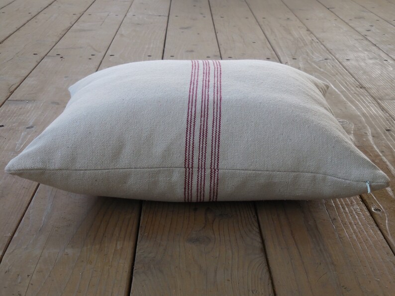 May include: A cream-colored pillow with a red stripe down the center. The pillow is made of a soft, textured fabric and has a zipper closure.