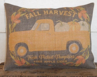 May include: Rectangular burlap pillow with a fall harvest theme. The pillow features an orange truck carrying pumpkins, with the words "FALL HARVEST" and "Fresh Picked Pumpkins FREE APPLE CIDER" printed on it. The pillow has a gray background.