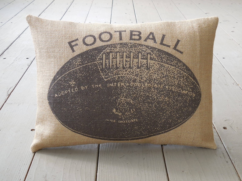 May include: A brown burlap pillow with the word "FOOTBALL" printed in black. The pillow features a vintage illustration of a football with the text "Adopted by the Intercollegiate Football Association" printed on the ball.