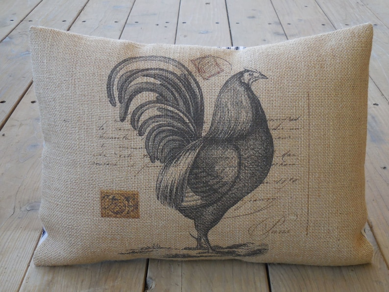 K&ouml;nnte beinhalten: Ein Kissen aus Jute mit einer Schwarz-Wei&szlig;-Illustration eines Hahns. Der Hahn steht auf einem braunen Hintergrund mit einem Design im Vintage-Stil.
