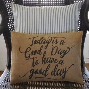 May include: A brown burlap cushion with the text "Today is a Good Day to have a good day" printed in black. The cushion is sitting on a black wicker chair with a white and black striped cushion behind it.