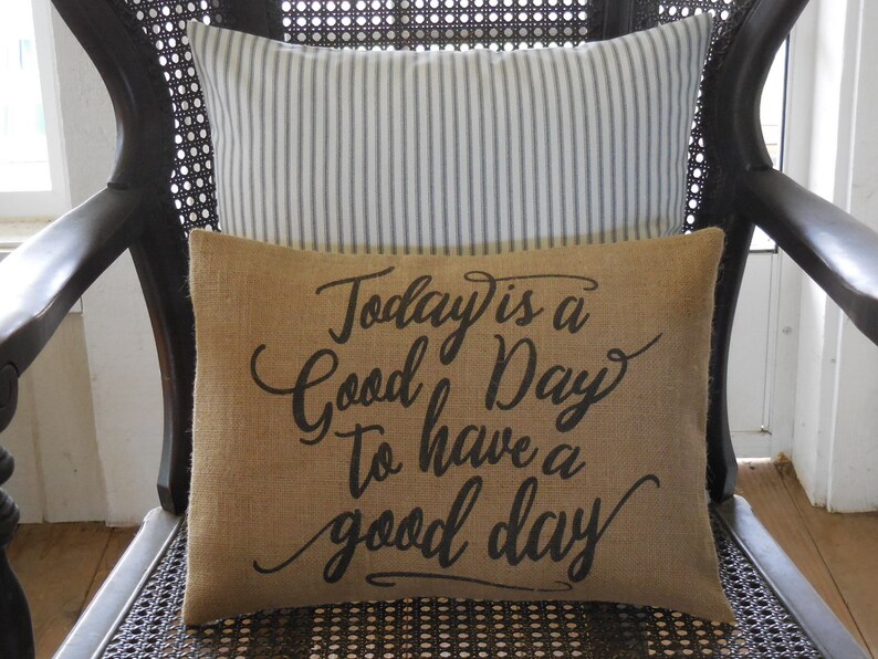 May include: A brown burlap cushion with the text "Today is a Good Day to have a good day" printed in black. The cushion is sitting on a black wicker chair with a white and black striped cushion behind it.