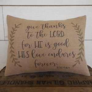 May include: A burlap pillow with a brown inscription that reads "give thanks to the LORD. for HE is good. HIS love endures forever. PSALM 104". The pillow has green leafy vines on each side.