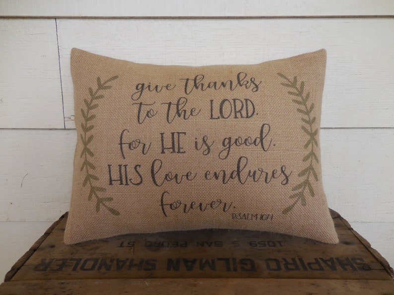 May include: A burlap pillow with a brown inscription that reads "give thanks to the LORD. for HE is good. HIS love endures forever. PSALM 104". The pillow has green leafy vines on each side.