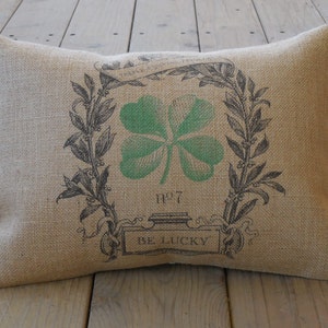 Puede incluir: Una almohada de arpillera con un diseño de trébol de cuatro hojas verde y el texto "Be Lucky".