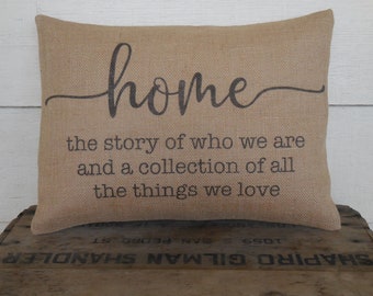 Może przedstawiać: Poduszka z juty z napisem "home" w ozdobnym, pochylonym piśmie. Poduszka zawiera również tekst "the story of who we are and a collection of all the things we love".