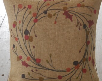 May include: A burlap pillow with a wreath design featuring brown branches, red berries, and green leaves.