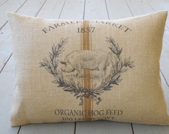 May include: A burlap pillow with a brown and white pig illustration and the text "Farmer Market 1857 Organic Hog Feed 100 lbs Net Wt."
