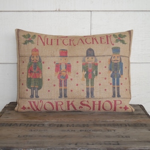 May include: A burlap pillow with a red and white printed design of four nutcracker figures and the text "Nutcracker Workshop".