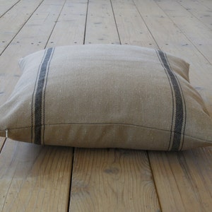 May include: A large, rectangular floor cushion with a beige linen fabric and black stripes. The cushion is lying on a wooden floor.