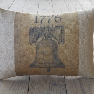 May include: A brown burlap pillow with a white border. The pillow has a black and white image of the Liberty Bell with the year 1776 printed above it.