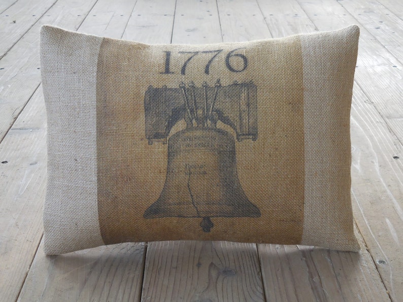May include: A brown burlap pillow with a white border. The pillow has a black and white image of the Liberty Bell with the year 1776 printed above it.