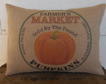 May include: A burlap pillow with a green circle design featuring the text "Farmer's Market", "Sold By The Pound", "Organic Produce", and "Pumpkins". A large orange pumpkin is in the center of the circle.