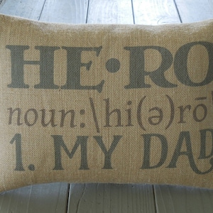 May include: A tan burlap pillow with the words "HERO" in dark gray, followed by "noun: /hi(ə)roʊ/" and "1. MY DAD." in a lighter gray.