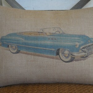May include: A blue vintage convertible car printed on a burlap pillow. The pillow is a light brown color and has a rough texture.