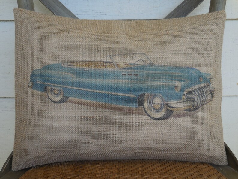 May include: A blue vintage convertible car printed on a burlap pillow. The pillow is a light brown color and has a rough texture.