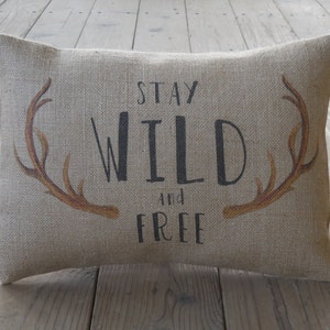 Puede incluir: Una almohada de arpillera beige con el texto "Stay Wild and Free" impreso en negro. La almohada tiene dos siluetas de astas marrones a cada lado del texto.