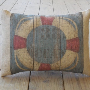 May include: A rectangular brown burlap pillow with a red, white, and blue life preserver design. The life preserver has a rope design and an anchor in the center.