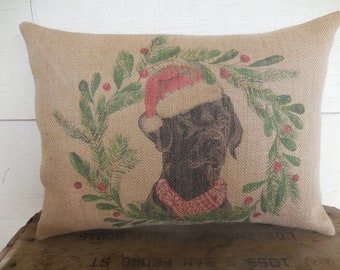 May include: A brown burlap pillow with a printed image of a black Labrador Retriever wearing a red Santa hat and a red and white scarf. The dog is surrounded by a green wreath with red berries.