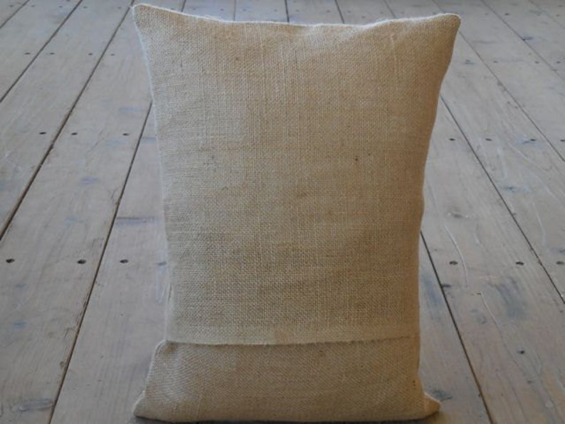 May include: A natural-colored burlap pillow with a simple design. The pillow is made of a coarse, woven fabric and has a slightly textured surface.