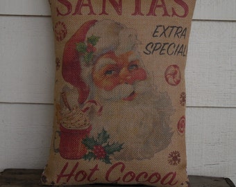 May include: A brown burlap pillow with a vintage illustration of Santa Claus holding a mug of hot cocoa. The pillow has the text "SANTA'S EXTRA SPECIAL HOT COCOA" printed on it.