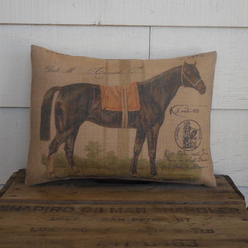 Op de afbeelding: Een bruin paard met een zadel en hoofdstel is gedrukt op een kussen van jute. Het kussen heeft een vintage uitstraling. De tekst "Est M. Agnes C" is op het kussen gedrukt.