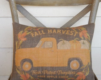 May include: A brown burlap pillow with a yellow pickup truck carrying pumpkins. The pillow has the text "Fall Harvest" and "Fresh Picked Pumpkins Free Apple Cider" printed on it.