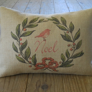 May include: A burlap pillow with a red bird and green leaves printed on it. The pillow has the word "Noel" printed in red on it.