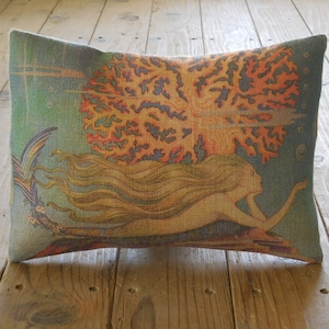 May include: A decorative pillow with a burlap-like texture features a colorful illustration of a mermaid swimming through a coral reef. The mermaid has long flowing hair and a tail with a fin. The coral reef is a vibrant orange and yellow with blue accents.