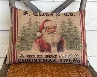 Może przedstawiać: Prostokątna poduszka z juty z ilustracją vintage Świętego Mikołaja w czerwonym stroju i czapce, z napisem "S. Claus & Co." i "Christmas Trees". Poduszka ma czerwone paski na krawędziach i jest umieszczona na drewnianym krześle.
