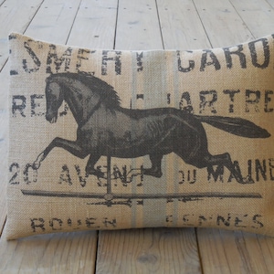 May include: A brown burlap pillow with a black and white image of a horse running on a carousel. The pillow has the text "Smerh Gardon", "Re", "Jartre", "20 Avenue du Maine", and "Rouen Gennes" printed on it.