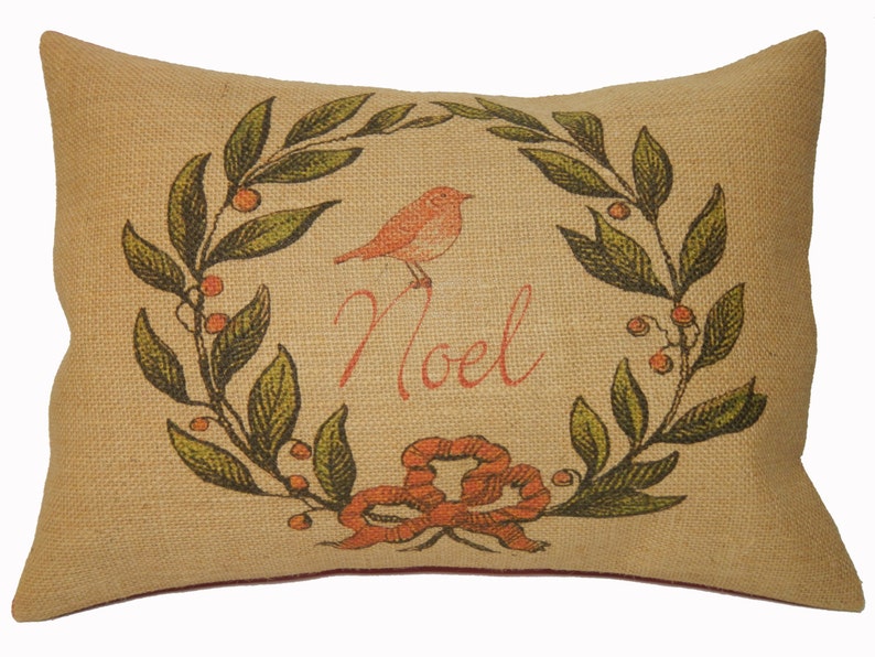 May include: A beige burlap pillow with a red bird and green leaves forming a wreath around the word "Noel".