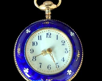 May include: A gold-toned pocket watch with a white face and blue enamel border. The watch features black numerals, gold hands, and decorative floral accents. The watch has a gold crown and a loop for a chain.