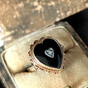 May include: A gold-toned ring with a heart-shaped black onyx center. A smaller heart-shaped design with a diamond is set in the center of the onyx. The ring is in a velvet-lined box.