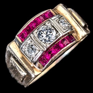 May include: A gold ring featuring a central row of three round clear stones set in white metal, flanked by two parallel rows of square-cut vibrant red stones. The band has a wide, polished gold finish with a structured, rectangular design.