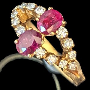 May include: A gold ring with two oval rubies and several round diamonds. The rubies are a deep red, and the diamonds are set in a gold band. The ring's design is detailed, with the gemstones arranged in a decorative pattern.